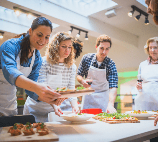 2 women and 1 man in cooking aprons placing platters of food on a benchtop